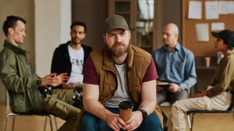 A focused veteran man holding a coffee cup sits in a group therapy session, surrounded by others engaging in discussion, fostering support and connection.