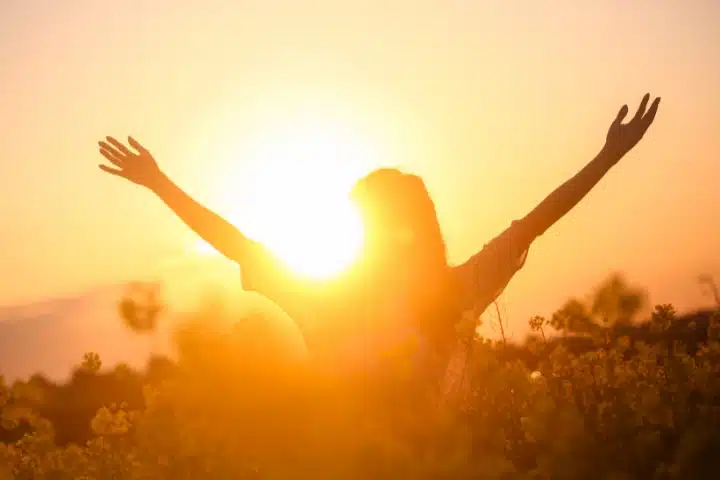 A silhouette of a woman raising her arms in joy, surrounded by golden light and flowers at sunset.