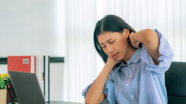 A young woman in a striped blue shirt sitting at a desk, holding her neck with both hands, appearing stressed or experiencing discomfort, with a laptop and books nearby.