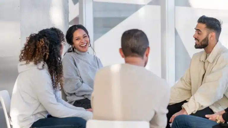 Group therapy session with diverse individuals sitting in a bright room, sharing and engaging in open, supportive conversation.