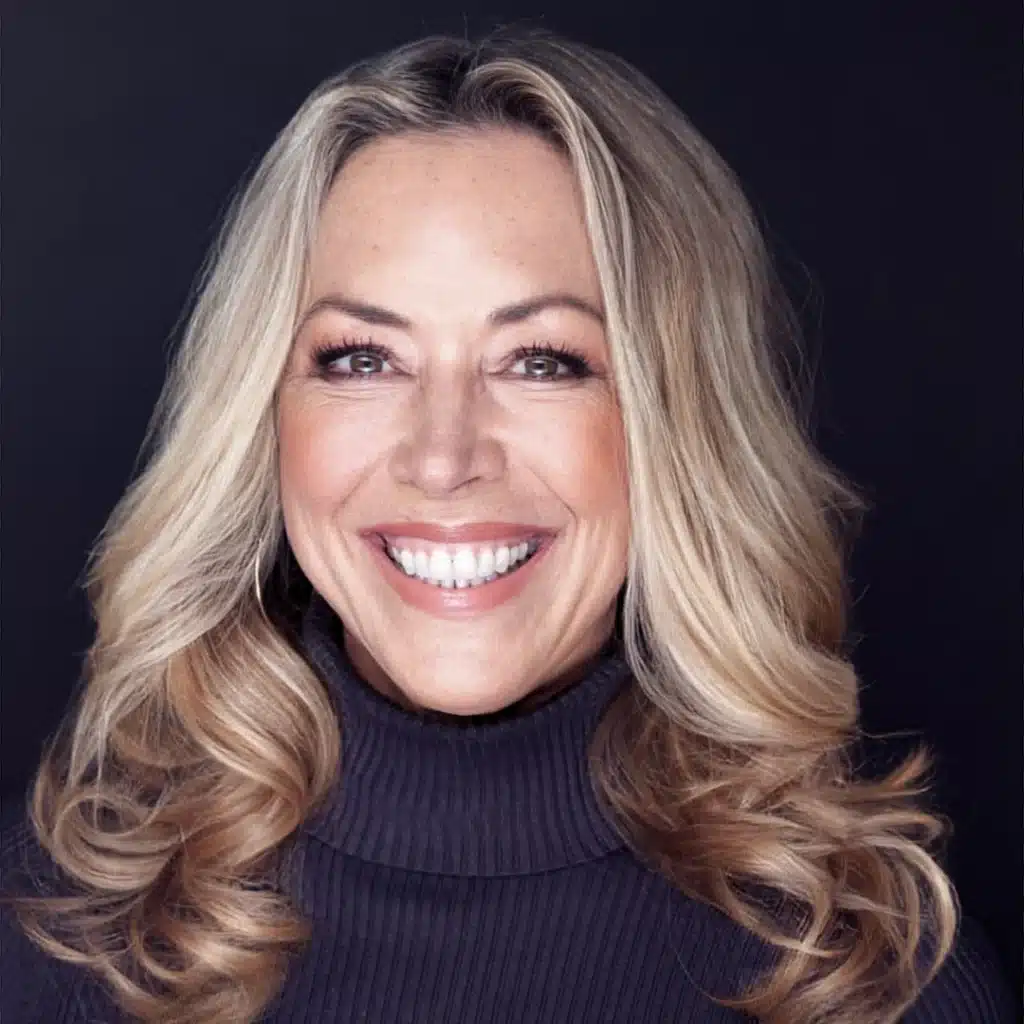 Smiling woman with long blonde hair wearing a dark turtleneck, posed against a solid dark background.