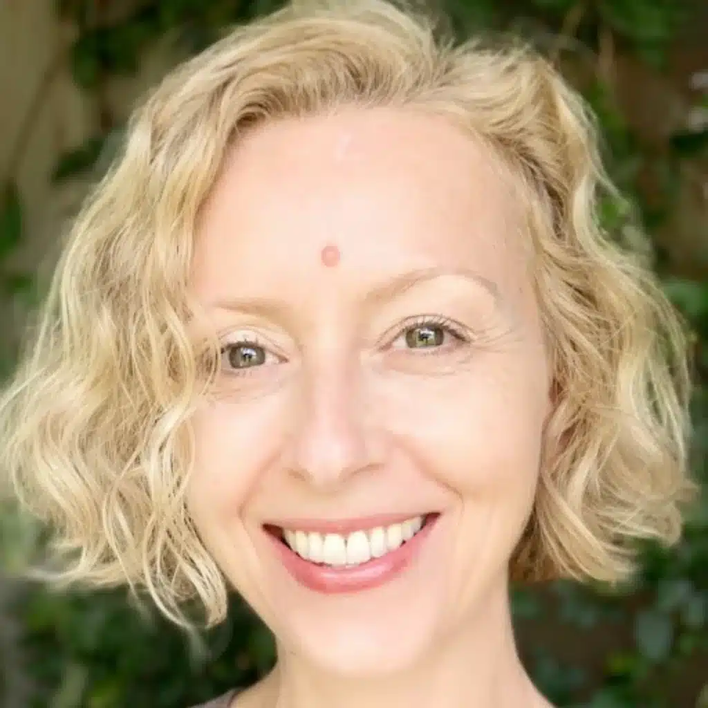 Smiling woman with short blonde hair and green eyes, standing in front of a leafy background.