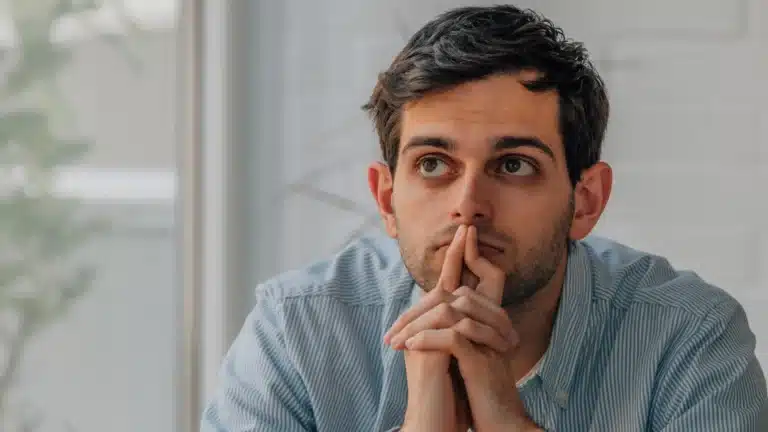 Man in a striped shirt resting his chin on clasped hands, gazing thoughtfully to the side, with a soft-focus background of a window and greenery.