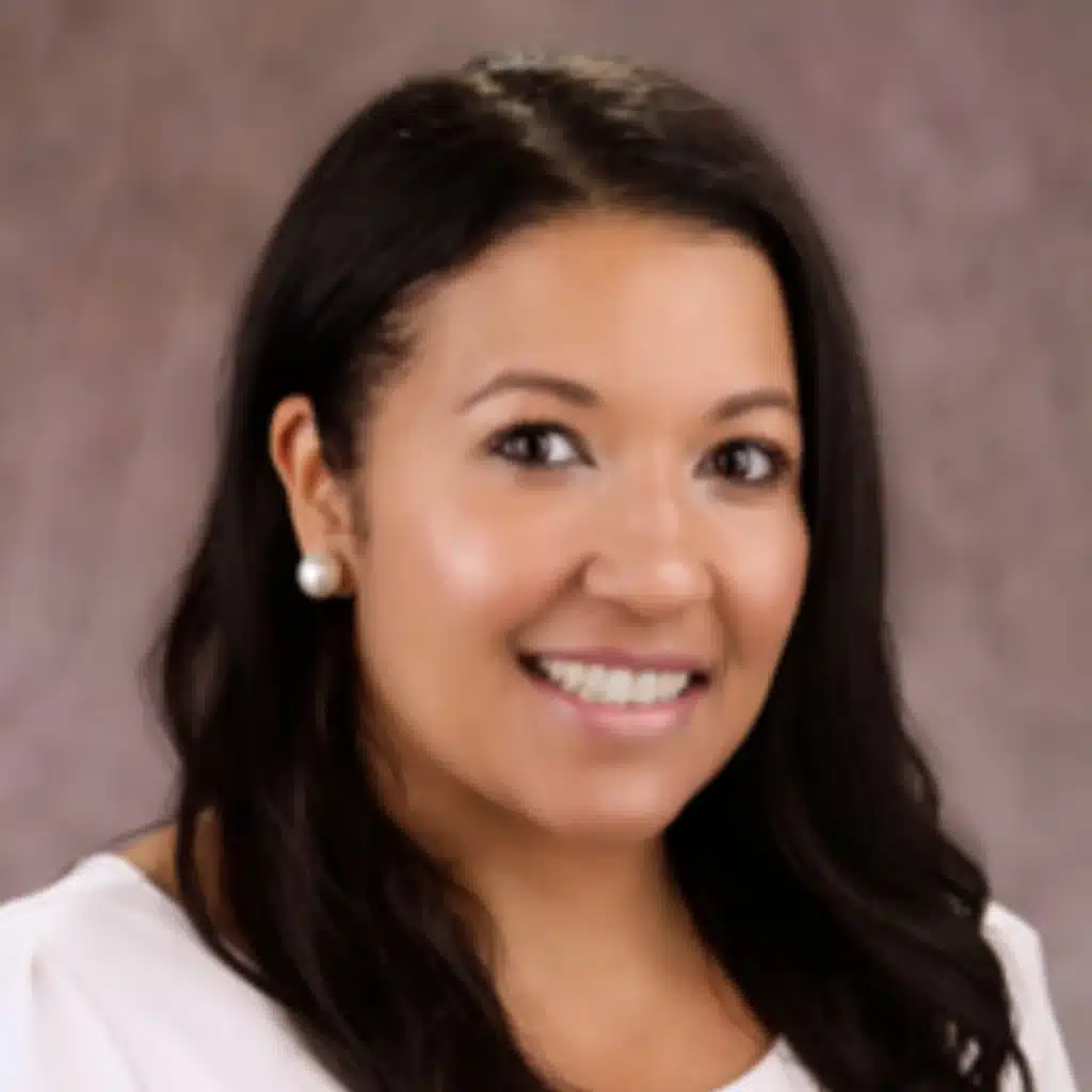 Smiling woman with long dark hair wearing a white blouse and pearl earrings, posed against a neutral background.