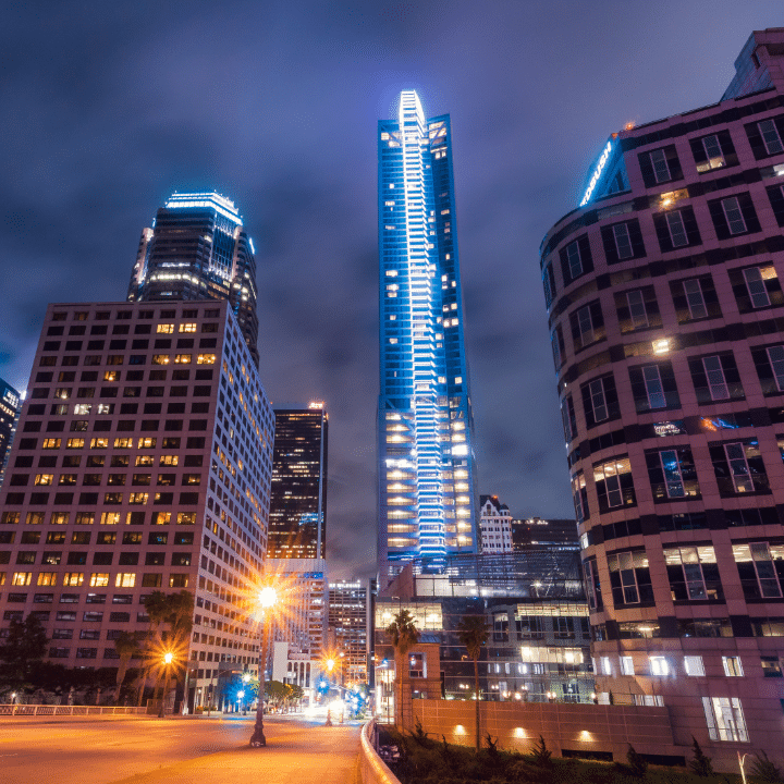 Night view of downtown Los Angeles featuring illuminated skyscrapers, including a prominent building with vertical blue lights, under a cloudy sky.