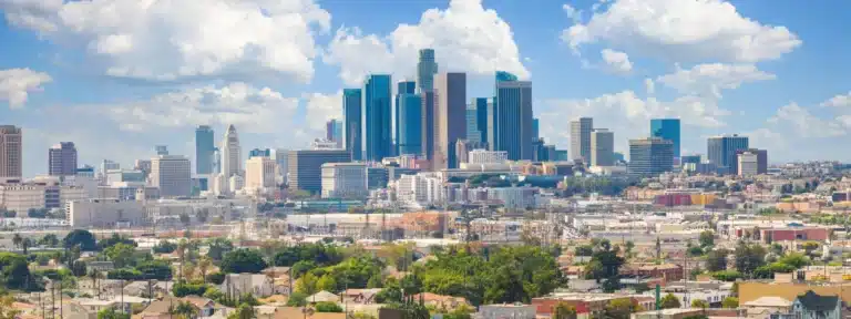 Panoramic view of downtown Los Angeles skyline with modern skyscrapers, residential areas in the foreground, and a bright blue sky with scattered clouds.