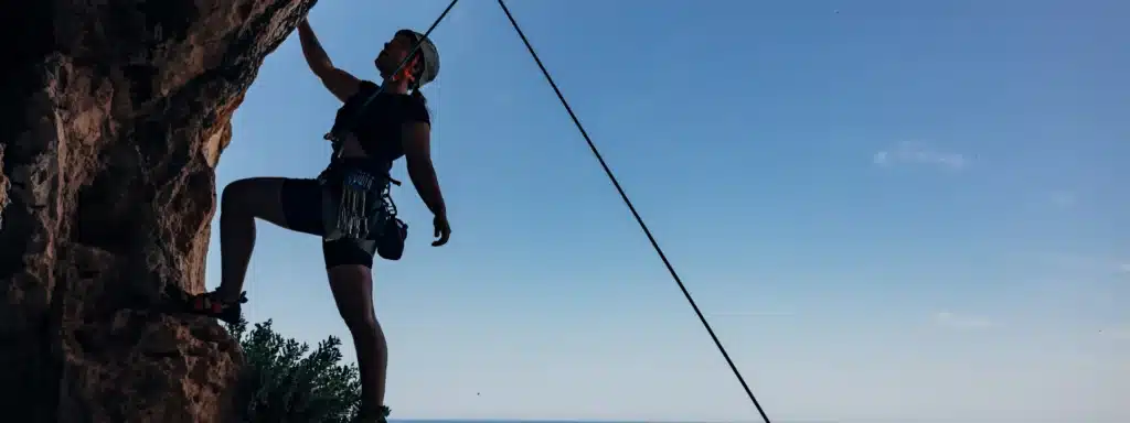 Person rock climbing with safety gear, silhouetted against a clear blue sky, symbolizing resilience, determination, and personal growth.