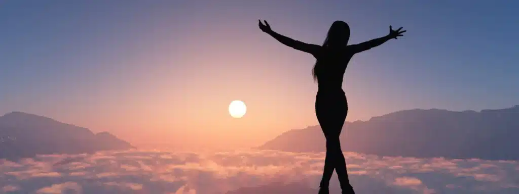 Silhouette of a person standing on a mountain, arms outstretched, facing a serene sunrise above a sea of clouds.