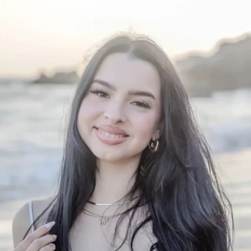 Smiling woman with long dark hair standing outdoors near a beach at sunset.