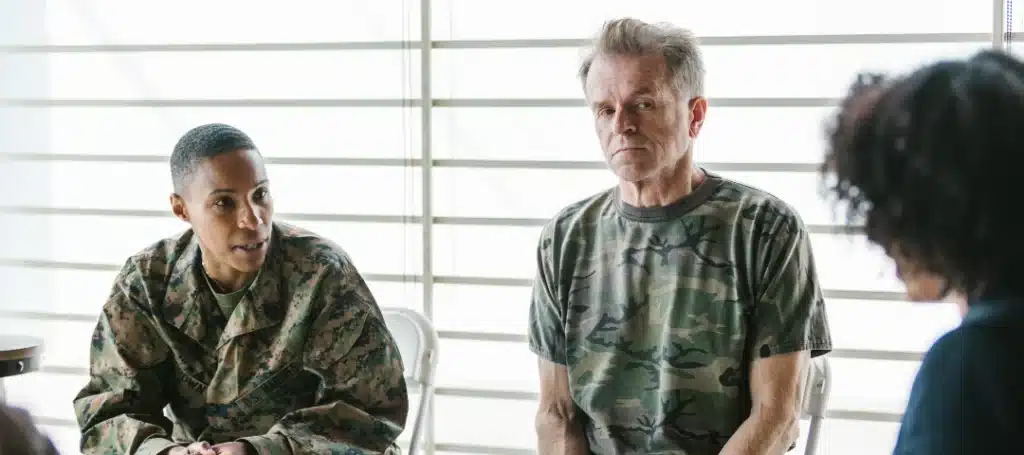 Two individuals in military uniforms engage in a thoughtful discussion during a group therapy session, seated in a well-lit room with a calm atmosphere.