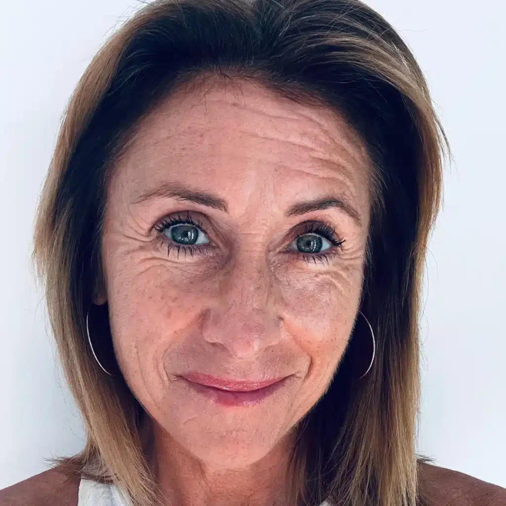 Smiling woman with light brown hair, wearing a white top and hoop earrings, posed against a plain background.