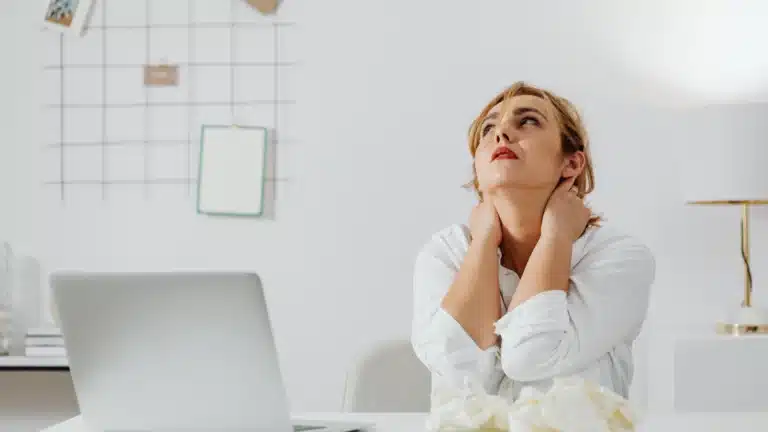 Woman sitting at a desk with a laptop, looking up with a tired expression, hands massaging her neck in a bright, minimalist workspace.