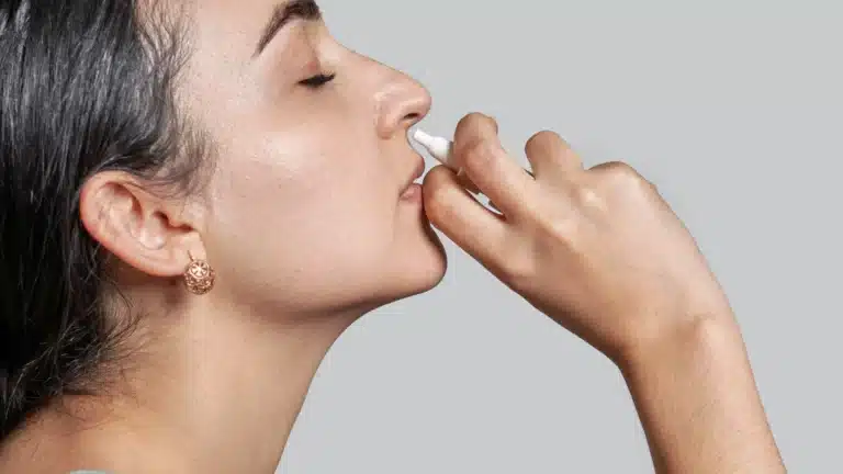 A close-up of a woman using a nasal spray, holding it near her nostril with a calm expression.