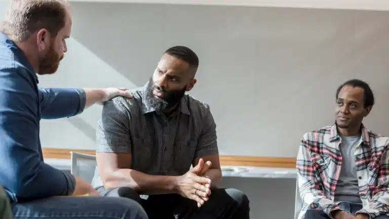 A support group setting with three men, one of whom is receiving reassurance as another places a hand on his shoulder.