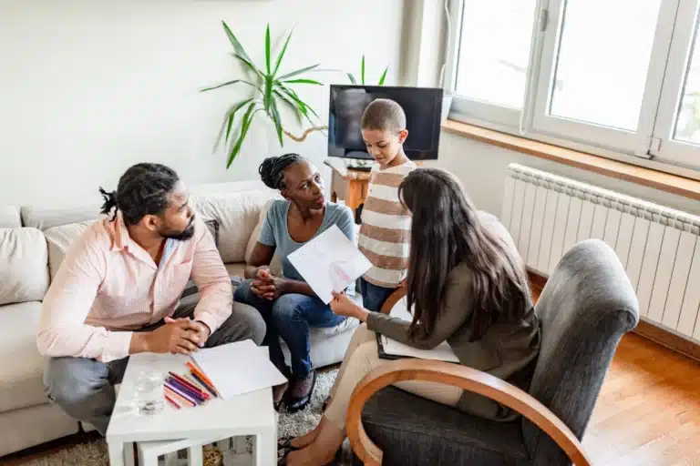 A therapist talking to a family in a home setting. A professional is holding a paper while discussing with a couple and a young boy.