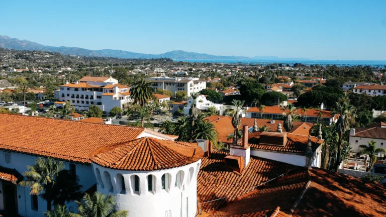 Aerial view of Santa Barbara, California, featuring red-tiled rooftops, Spanish-style architecture, palm trees, and the Pacific Ocean in the background.