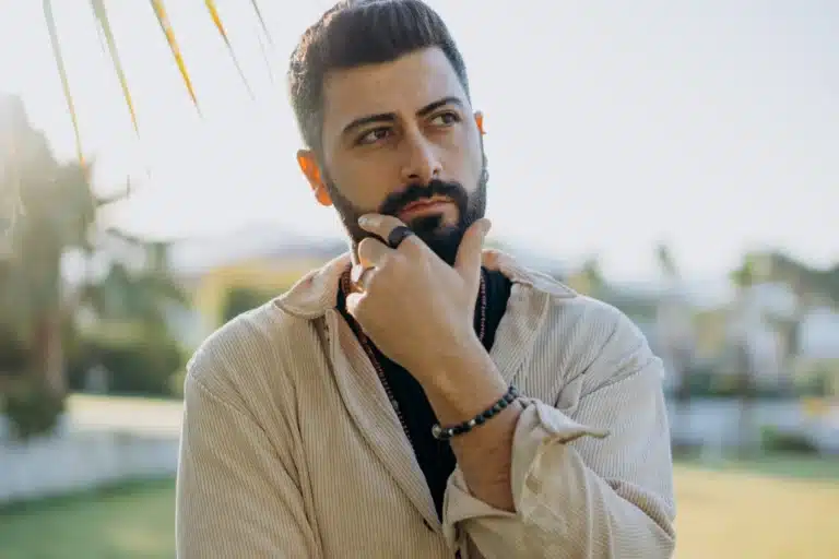 Bearded man wearing a beige shirt, standing outdoors in a sunny setting, thoughtfully resting his hand on his chin with palm trees in the background.