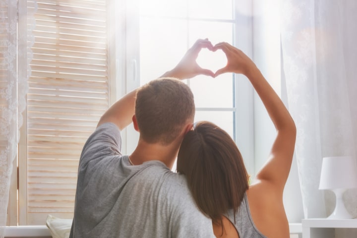 A couple sits together near a window, forming a heart shape with their hands as sunlight streams through the curtains.