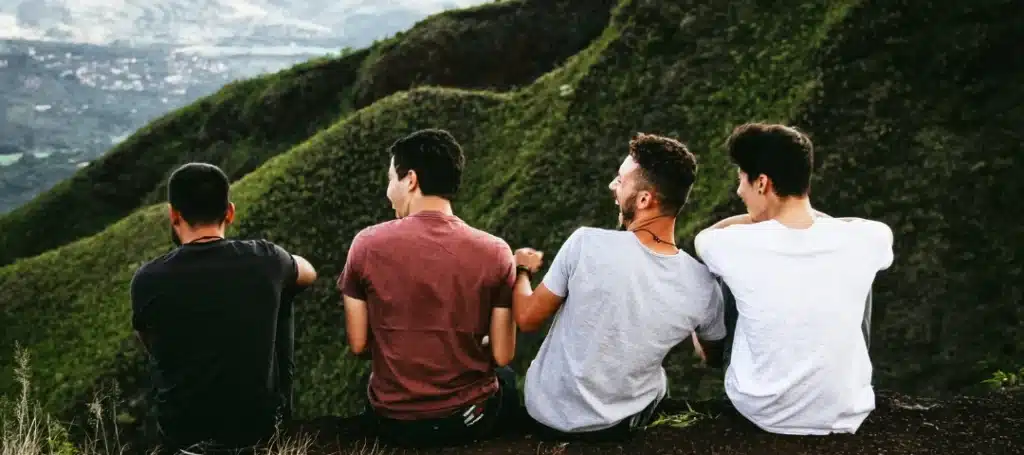 Four friends sitting on a mountain ledge, overlooking green hills and valleys while laughing together.