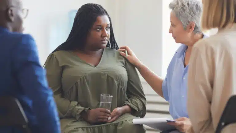 Group therapy session with a diverse group of individuals seated in a circle, with an older woman offering support to a younger woman holding a glass of water.