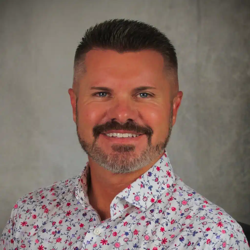 A smiling man with neatly styled hair and a trimmed beard, wearing a white shirt with a colorful floral pattern, posing against a neutral background.