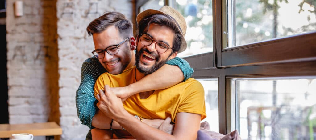 A happy same-sex couple shares a warm embrace in a cozy cafe, laughing together as they enjoy each other’s company.