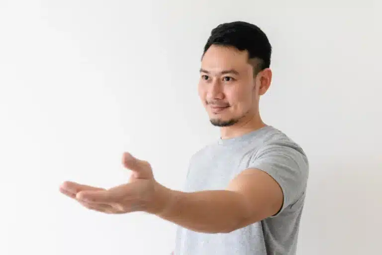 A man wearing a gray t-shirt extending his hand for a handshake with a welcoming smile against a white background.