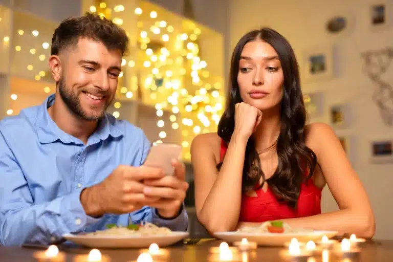 A man in a blue shirt smiling while looking at his phone, while a woman in a red dress rests her chin on her hand, appearing displeased.