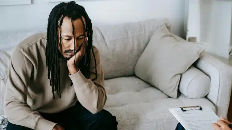 Man with dreadlocks sitting on a couch, looking distressed. He rests his head on his hand while a clipboard with notes is visible in the foreground, indicating a therapy session or deep contemplation.