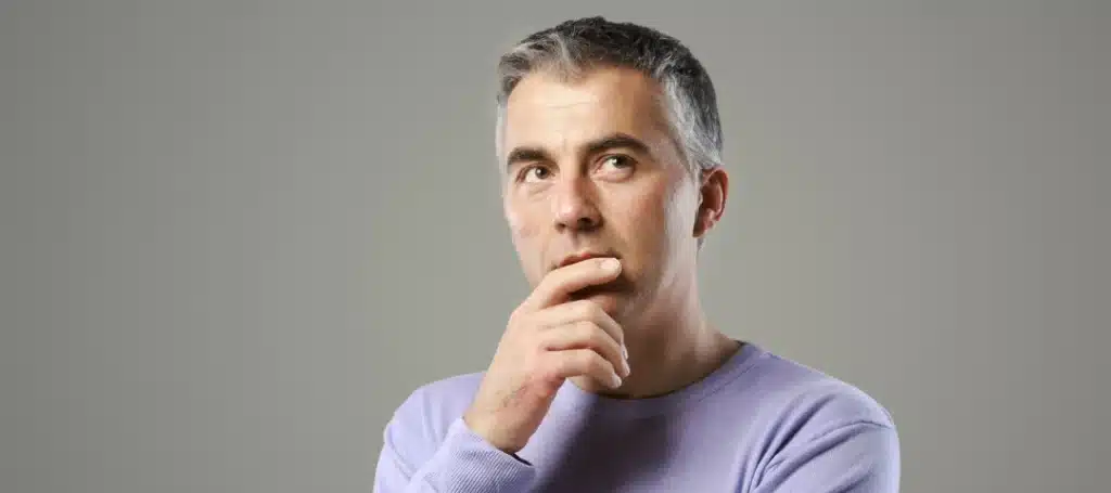 Middle-aged man with gray hair wearing a light purple sweater, looking thoughtful with his hand on his chin against a plain gray background.