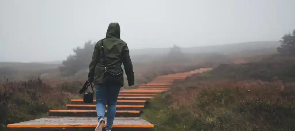 Person in a hooded jacket walking up a misty wooden path in a foggy landscape.