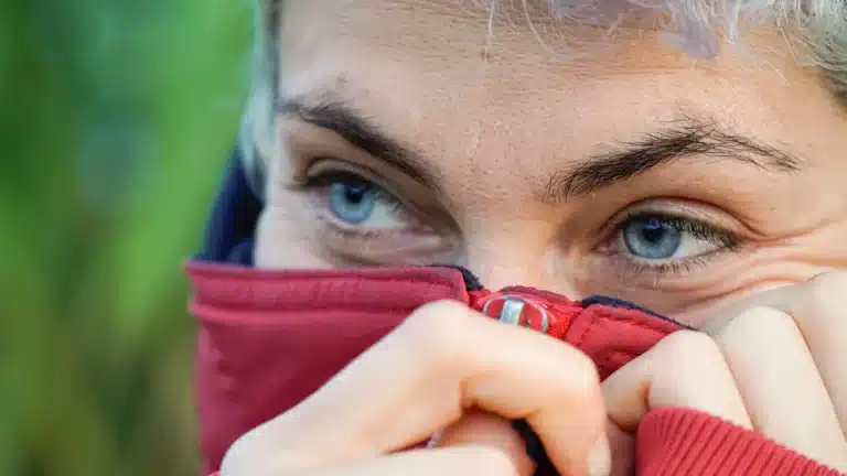 A close-up of a person with striking blue eyes pulling their red jacket up to cover their face.