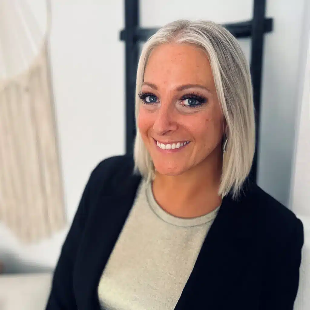 A professional headshot of a smiling woman with short blonde hair, wearing a black blazer and light-colored top, seated indoors.