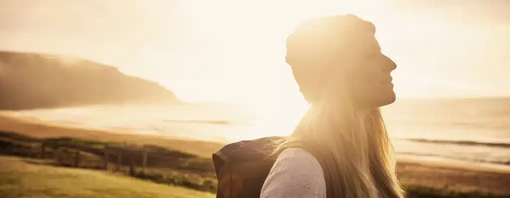 Side profile of a woman enjoying a serene beach view at sunrise or sunset, wearing a beanie and carrying a backpack.