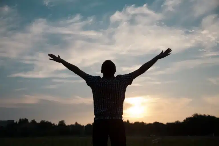 Silhouette of a man with arms raised, standing in an open field at sunset under a partly cloudy sky.