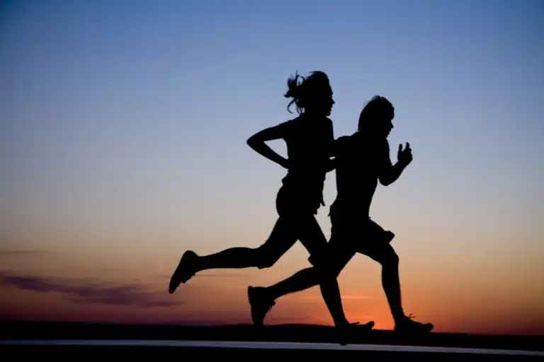 Silhouette of two people jogging side by side at sunset, with a vivid sky and horizon in the background.