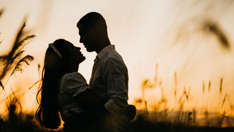 A couple stands in a field, gazing at each other as the sun sets behind them, casting a warm glow over the scene.