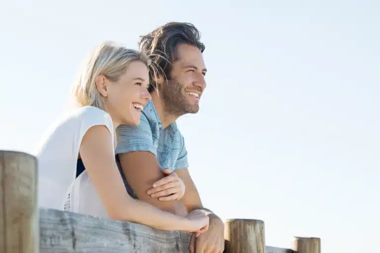 Smiling couple leaning on a wooden railing outdoors, looking into the distance under a bright sky.