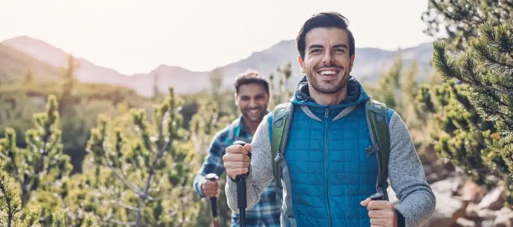 Two men hiking on a mountain trail, wearing backpacks and holding trekking poles, surrounded by trees and sunlight in a scenic outdoor setting.