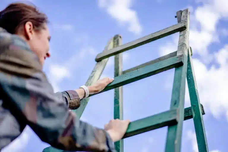 Woman climbing a wooden ladder, reaching upward under a bright blue sky with scattered clouds.