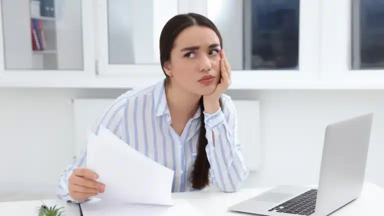 Woman sitting at a desk with a laptop, holding papers and resting her head on her hand, looking frustrated in a bright office setting.