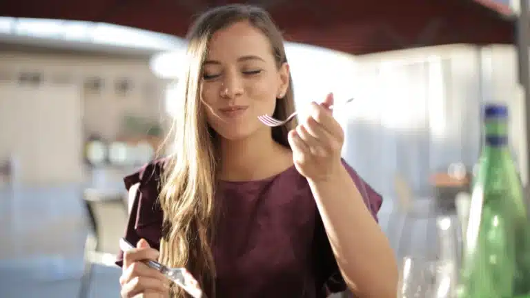 Woman sitting at an outdoor table, enjoying a meal with a fork in hand, smiling contentedly, with sunlight and a blurred background.