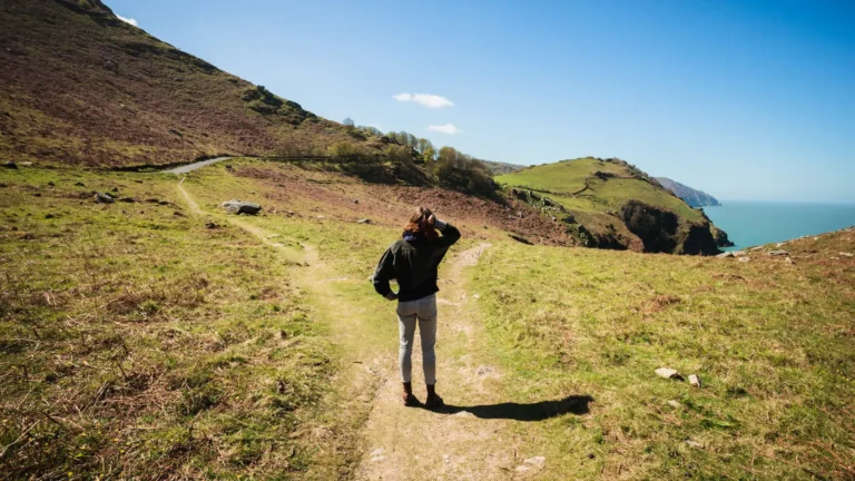 Woman standing on a dirt path overlooking rolling green hills and the ocean in the distance.