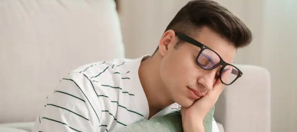 Young man wearing glasses and a striped shirt, asleep on a couch with his head resting on a pillow.