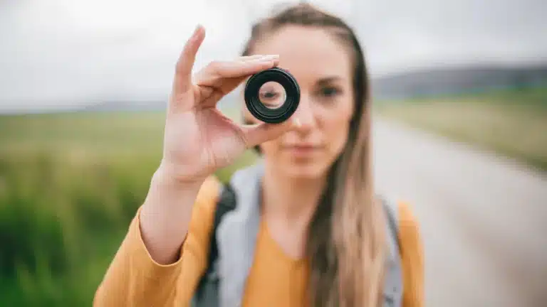 Young woman holding a camera lens up to her eye, focusing through it, with a blurred outdoor background of a rural path and green fields.