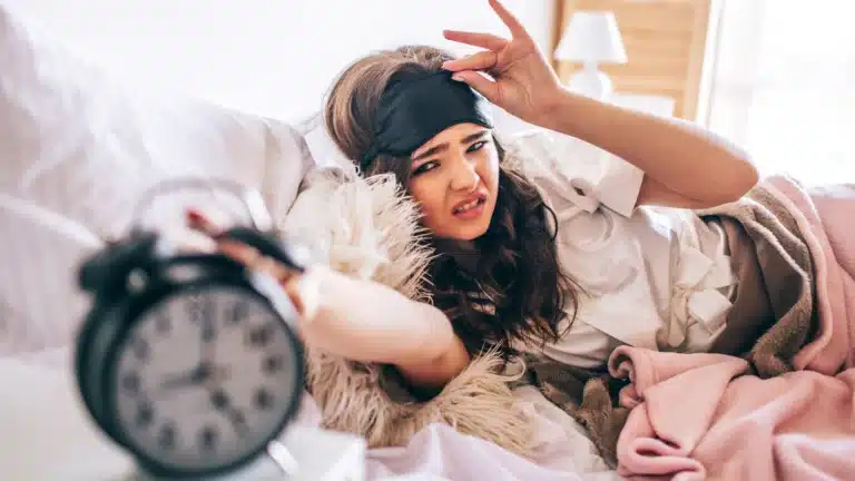 Young woman in bed reaching for an alarm clock with a frustrated expression, wearing a black sleep mask.