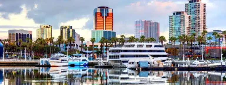 Scenic view of downtown Long Beach, California, featuring yachts docked in the marina, palm trees, and modern high-rise buildings reflecting in the water.